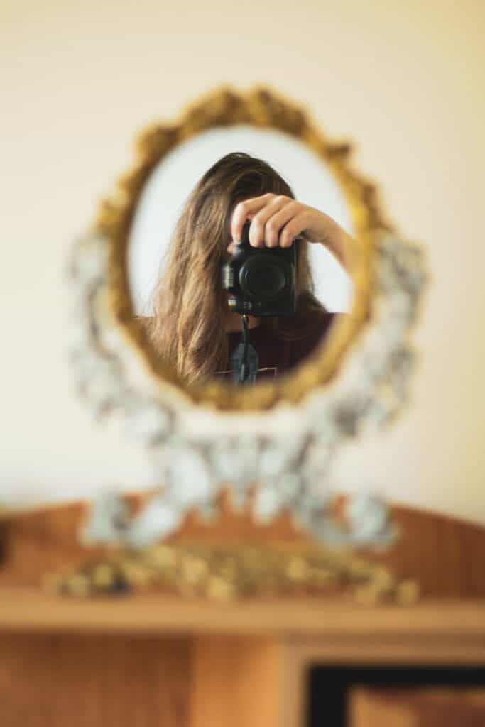 pexels-photo-15657120-15657120 A woman takes a selfie with a DSLR camera in an ornate oval mirror indoors.